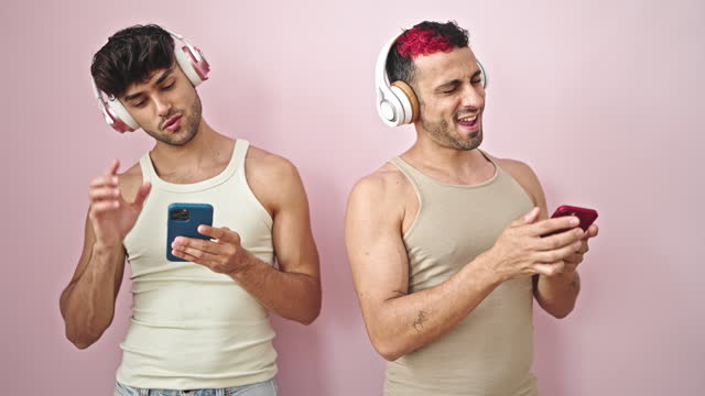 Two men couple listening to music dancing together over isolated pink background