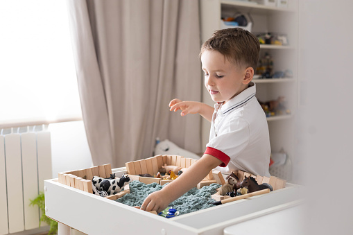 Cute baby boy playing sensory box kinetic sand table with farm animals. Montessori material.