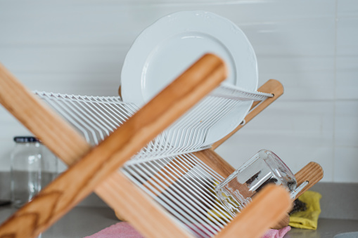 A white plate placed on a wooden dishrack next to a sink. Concept of washing the dishes by hand and leaving them to dry. Household chores.