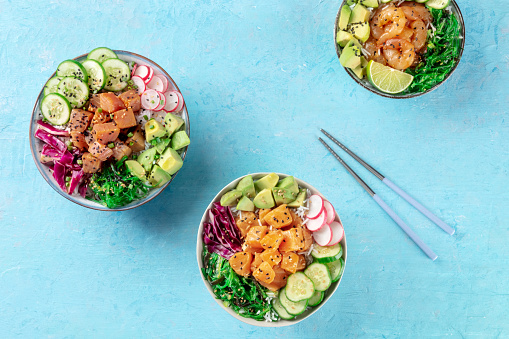 Poke bowl variety. Healthy Hawaiian dish with salmon, tuna, and shrimps, overhead flat lay shot on a blue background, with chopsticks