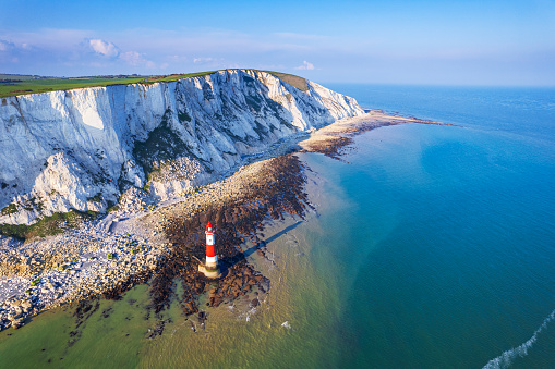 Rear view image depicting a senior man in his 70s sitting on a bench leading down to the iconic Seven Sisters cliffs on the coastline of East Sussex, UK. The man is wearing casual clothing - blue denim jeans, a red checked shirt, navy blue gilet. The man is having a rest from hiking and his hiking pole is leaning up against the bench.