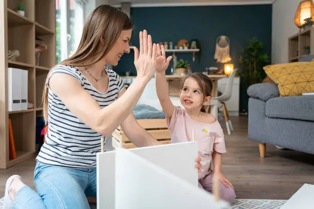 Young Caucasian woman interchange high-five with her toddler daughter while together assembling new furniture