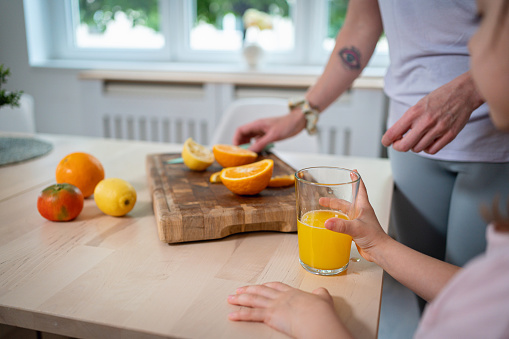 Unrecognizable young Caucasian mother cutting orange, while her daughter drinking juice