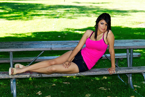 Young Latina Woman Teen Reclining On Park Bench In Shorts And Pink Top With Grass In Background