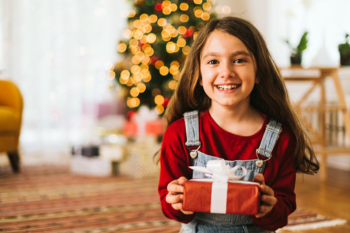 Smiling funny child (kid, girl) in Santa red hat holding Christmas gift in hand. Christmas concept.