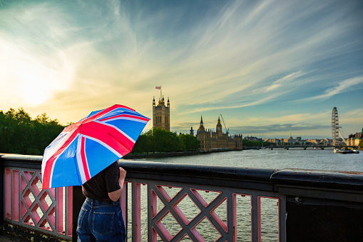 Color image depicting the rear view of an unrecognisable woman holding an umbrella with the design of the British union flag on it. It is a sunny day and the woman is using the umbrella for protection from the sun. The woman is looking at the view from Lambeth Bridge towards the Houses of Parliament and Big Ben in London.