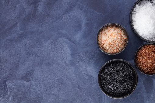 Different kinds of salts in a dark bowls, top view. Composition with various salt in a bowls on a blue background. Sea salt, cooking salt, Himalayan and black salt. Copy space.