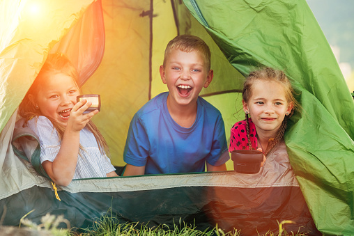 Little girls sisters and brother boy sitting inside green camp tent drinking tea and cheerfully laughing and looking into camera. Careless childhood, family values and outdoor activities concept photo
