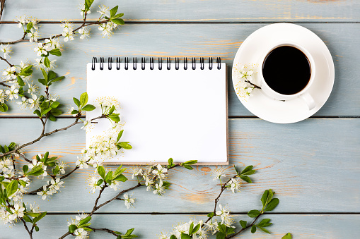 Artistic composition with opened blank notepad, cup of black coffee, branch of white cherry blossoms on blue wooden table. Festive office desktop concept. Morning coffee cup.