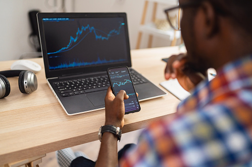 Over the shoulder view of unrecognizable African-American male trader, follow the graph of the stock market, while working from his modern home office