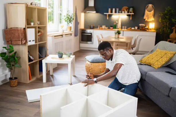 Young African-American man using electric screwdriver while  assembling new furniture at his modern apartment