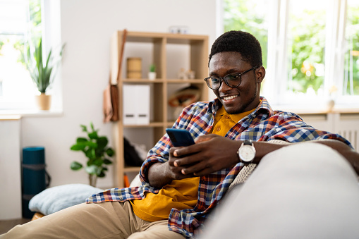 Young man of African-American ethnicity, sitting on the sofa while using mobile phone