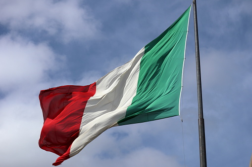 Flag of Italy blowing in bright Mediterranean sun on a flagpole standing in Italian blue sky