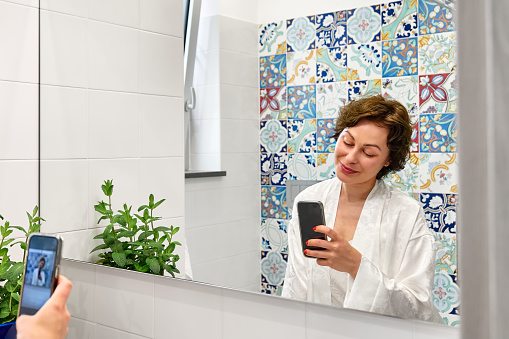Woman in white silk bathrobe smiling and using smartphone for selfie, chatting and blogging in front of mirror in bathroom in the morning.