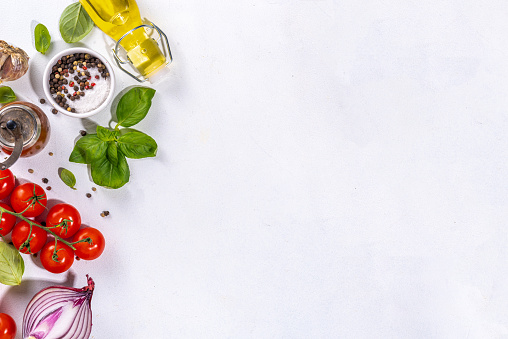 White cooking background with useful cooking italian Mediterranean ingredients - tomatoes, basil leaves, greens, olive oil, salt, pepper, garlic, flat lay white table top view copy space