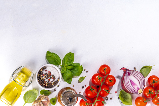 White cooking background with useful cooking italian Mediterranean ingredients - tomatoes, basil leaves, greens, olive oil, salt, pepper, garlic, flat lay white table top view copy space
