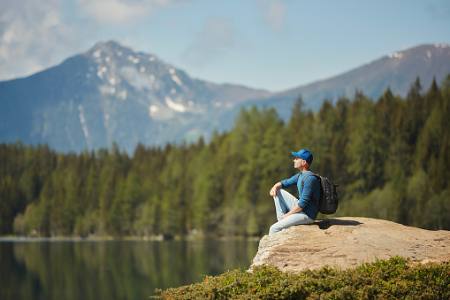 Man with backpack sitting on rock and enjoying scenic view of mountain lake. Young solo traveler in the Alps.