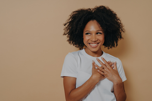 Happy African woman in white t-shirt posing with a smile, looking away, expressing gratitude with hands on chest, symbolizing sincere feelings and love.