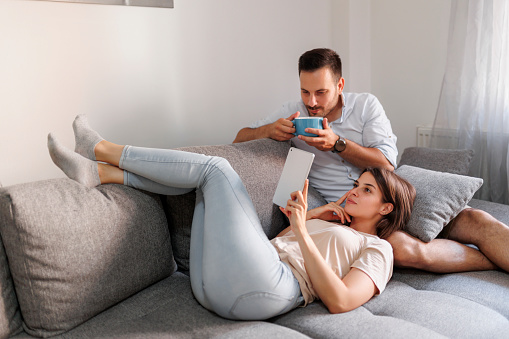 Beautiful young couple in love relaxing at home, surfing the Internet using tablet computer and drinking coffee in the morning