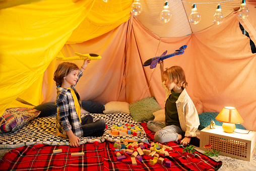 Two young guys with longer hair are having fun together in a comfy indoor tent, there are pillows behind them and blankets everywhere. Portrait