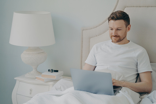 Cheerful young man, dressed in casual pajamas, sits in bed in the morning. Working remotely on a laptop while enjoying the comfort of his cozy bed. Embracing the freelance lifestyle.