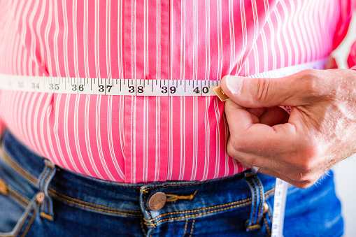 Close up color image depicting a senior man measuring his waistline with a tape measure.