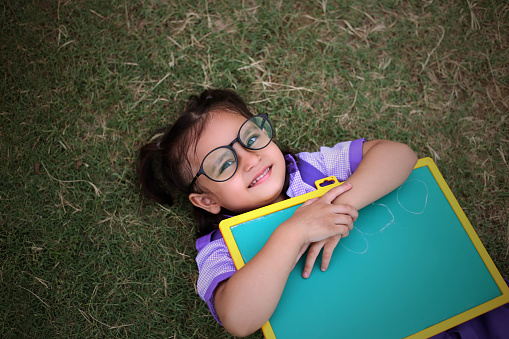 Smiling cute small girl with writing slate, laying on green grass outdoors.