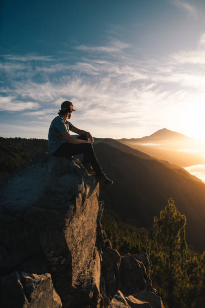 istock Young man with cap sitting on a rock observing the Teide peak at sunset during his touristic travel through the Canary Island of Tenerife. 1496433468