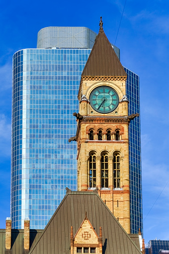 Toronto, Canada - May 13, 2023: Old City Hall. View of a striking clock tower on a building. Standing behind it is a sleek, tall blue building with a glass facade; no people are in the scene.