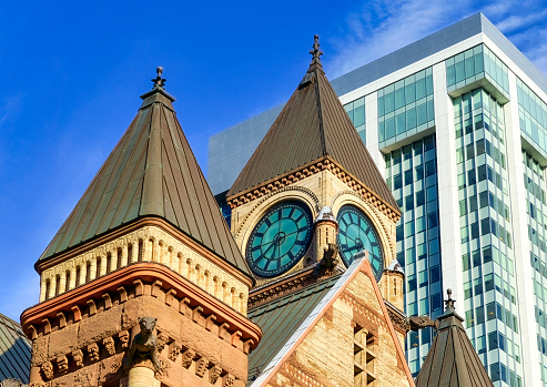 Toronto, Canada - May 13, 2023: The upper section of a clock tower building, accentuated against the backdrop of a tall building and a clear blue sky. No people are in the scene.