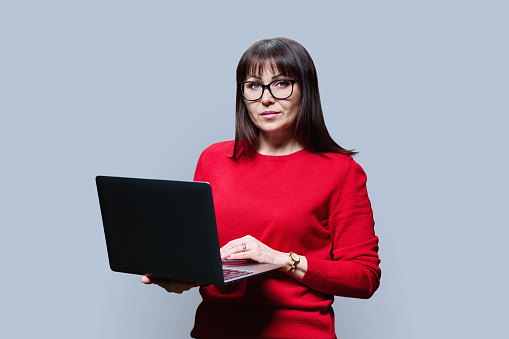 Serious mature woman using laptop looking at camera on grey studio background. Confident middle aged female in casual red jumper. Work, staff, freelancing, technology lifestyle leisure concept