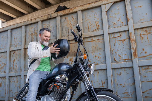 A mature man wearing a light green t-shirt, a beige jacket, and gray jeans on a Bobber Motorcycle. He holds a helmet in his hands. In the background - a gray and rusted metal container is under a large bridge.