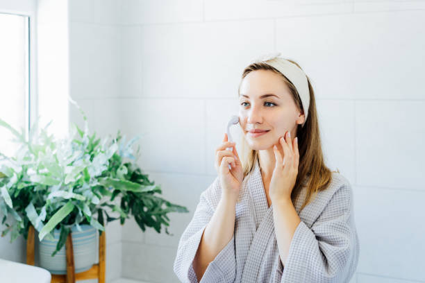 istock Young woman in bathrobe looking in mirror and making face massage with jade roller in the bathroom. The concept of natural procedures for skin care. Home Beauty self-care treatment. Selective focus. 1496439114