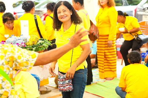 Group of yellow dressed thai women at Public local traditional culture event organized by local government near historical grounds of Wat Wihan Thong Historical Site near Nan river and is local way of keeping history and traditions alive. Women are packaging fresh made banana caramel or are customers to get fresh caramal. One woman is smiling into camera
