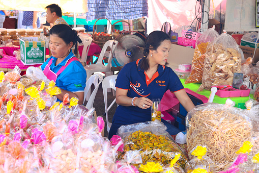 Working thai food vendor women at their market stall at Public local traditional culture event organized by local government near historical grounds of Wat Wihan Thong Historical Site near Nan river and is local way of keeping history and traditions alive. Women are selling several types of dried thai food. They are sitting behind their market stall