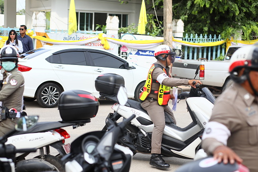 Thai traffic cop  on motorcycle is standing in street of Phitsanulok. Cop is wearing helmet and is overviewing and securing the traffic and  street. Scene is around a local public health and cycling event for thai people organized by provincial government