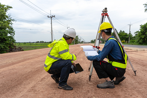 Asian engineer holding tablet to inspect work, with foreman using telescope to check pin against road plan, telescope and radio to control road construction, against road background under construction