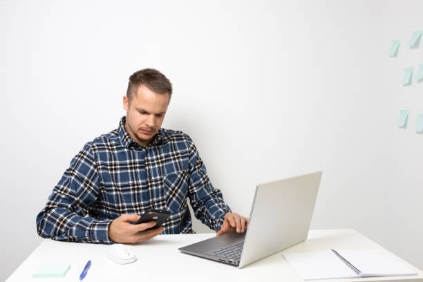 Portrait of a young man in a home office. Uses a phone and uses a laptop. Photo Portrait of a young man in a home office. Uses a phone and uses a laptop. Photo ERP for School stock pictures, royalty-free photos & images