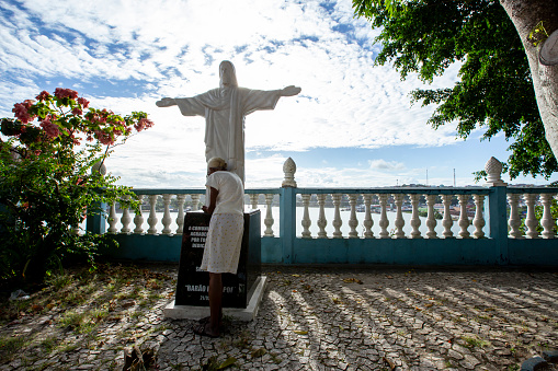Ilhéus, Bahia, Brazil – December, 01, 2016: old senior citizen lady praying at a Christ marble statue in silhouette at the sunset light.