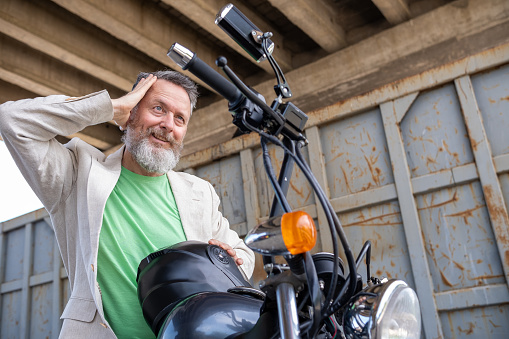 A mature man wearing a light green t-shirt, a beige jacket, and gray jeans on a Bobber Motorcycle. He fixes his hair and looks in the rearview mirror.  In the background - a gray and rusted metal container is under a large bridge.  He is looking back.