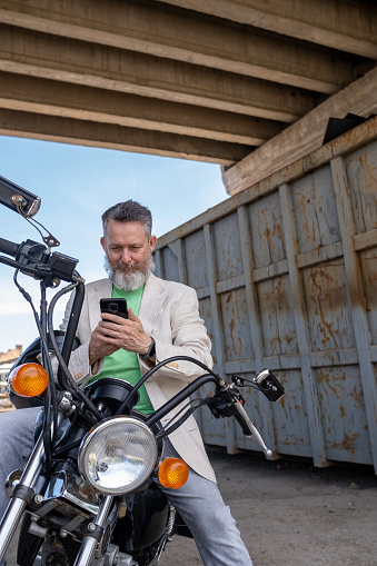 A mature man wearing a light green t-shirt, a beige jacket, and gray jeans on a Bobber Motorcycle. He looks at his phone with an interesting expression. In the background - a gray and rusted metal container is under a large bridge.