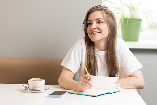 A young and attractive Caucasian girl is writing a book in a notebook while sitting in a cafe.