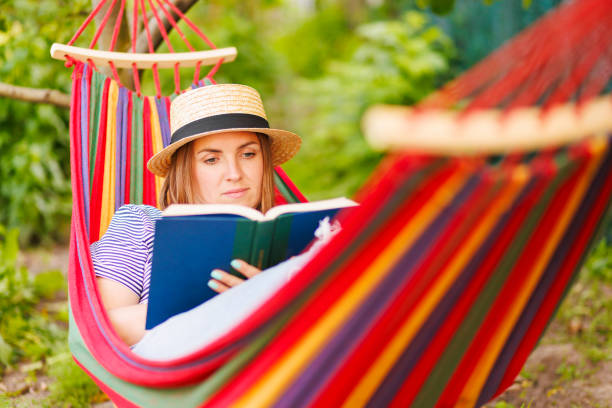istock Young woman reading book while lying in comfortable hammock at green garden 1496457666