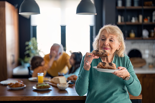 Happy satisfied senior woman enjoying smelling fresh home baked pastries in the kitchen at home standing in front of kitchen table. Senior man playing with kids in the blurred background.