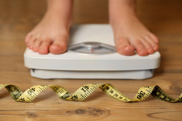 Overweight girl using scales near measuring tape on wooden floor, selective focus stock photo