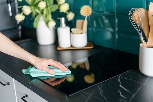cropped shot of female hand with microfiber rag cleaning glass ceramic electrical hob on marble countertop in stylish kitchen, housework concept