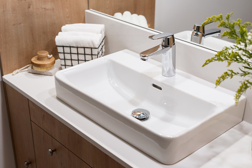 detail in interior of bathroom, modern ceramic wash basin with chrome water faucet, clean towels in metal basket and wooden massage brush