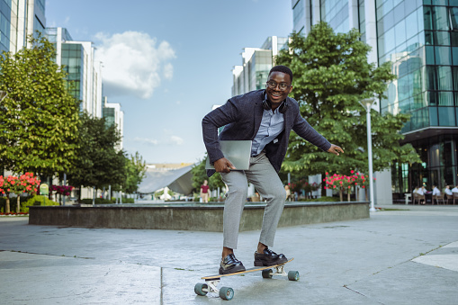Handsome African American young modern businessman holding laptop and riding a longboard