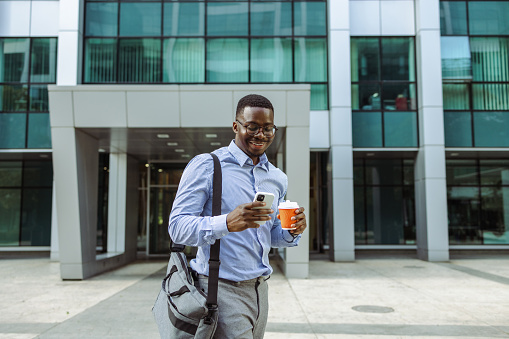 Young African-American businessman using a smart phone on the street. He is holding coffee to go and checking smart phone