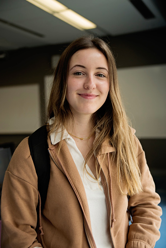 Portrait of young woman college student in classroom. She has long blond hair and is looking at the camera with a soft smile. Vertical waist up indoors shot with copy space. This was taken in Quebec, Canada.